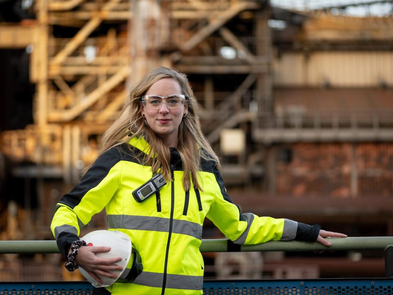 Frau in Sicherheitskleidung hält einen Helm, vor einer industriellen Kulisse. Julia Zoonz Arbeitssicherheit